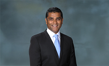 The image features a man wearing a suit with a tie, standing against a background with a blurred photograph of another person on the wall.