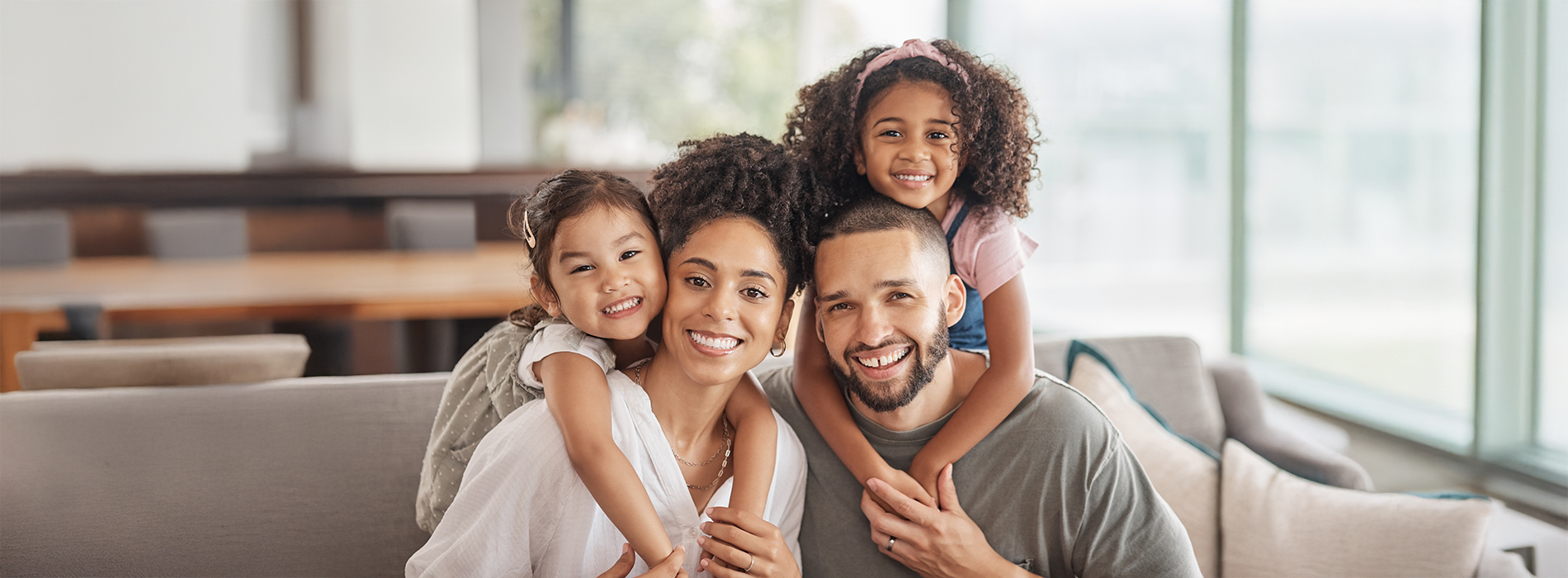 The image shows a family of four people, including two adults and two children, smiling together in an indoor setting with modern furniture.